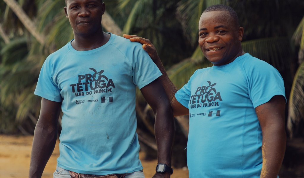 Two locals in light blue Pro Tetuga T-shirts and shorts smile at us on Príncipe Island beach.