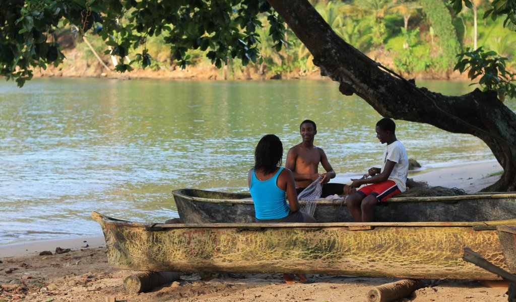 Three locals sitting in wooden boats on the beach by the sea on Príncipe Island.