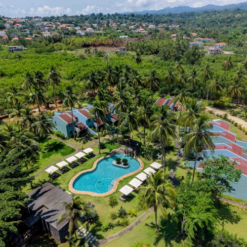 Aerial view of the Omali pool area and rooms surrounded by palm trees, with the town of São Tomé in the background