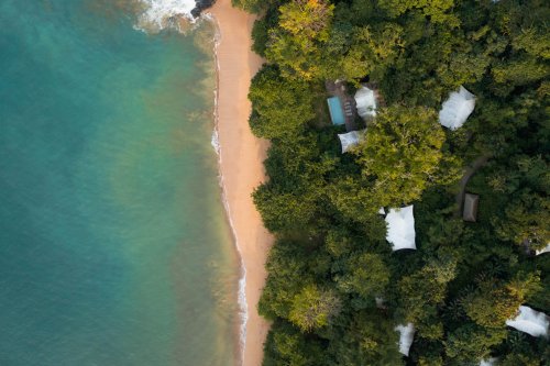 Vista aérea das tenda-vilas no Sundy Praia, quase escondidas pela selva, com a praia e a água cristalina à frente.