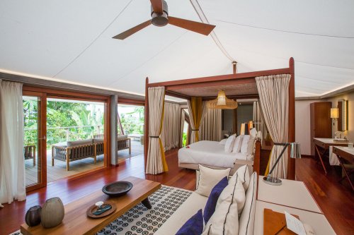 Bedroom with elegant four-poster bed, artistic rug, seating area, and wooden table in the foreground at Sundy Praia tent villa.