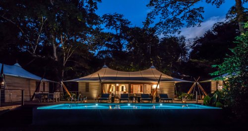 Exterior view of the tent villa at Sundy Praia by night, with the illuminated bedroom in the background and the pool in the foreground.