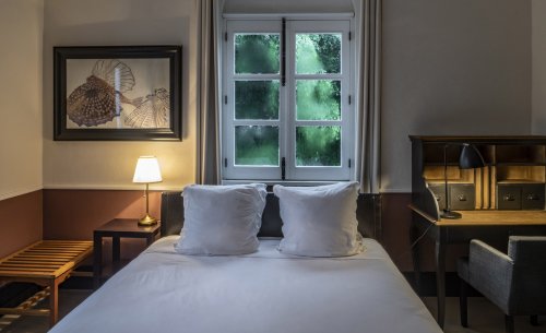 Cozy bedroom at Roça Sundy featuring a bed, a desk, a window with greenery view, and soft lighting