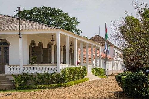 Vista exterior da entrada da Roça Sundy com pilares brancos, janelas arqueadas e vegetação exuberante