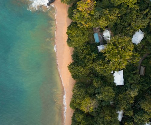 Aerial view of the tent villas at Sundy Praia, nearly hidden by the rainforest, with the beach and crystal-clear water in front.