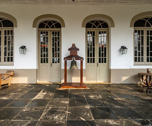 Entrance of Roça Sundy with colonial-style doors, a bell, and wooden chairs on the porch
