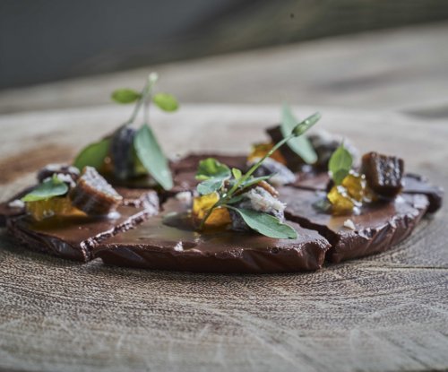 Close-up de uma elegante sobremesa de chocolate decorada com ervas e frutas, servida num prato de madeira no restaurante OKA Sundy
