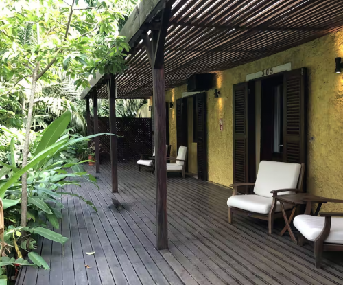 Varanda sombreada junto aos Quartos Jardim, deck em madeira com assentos entre vegetação tropical.