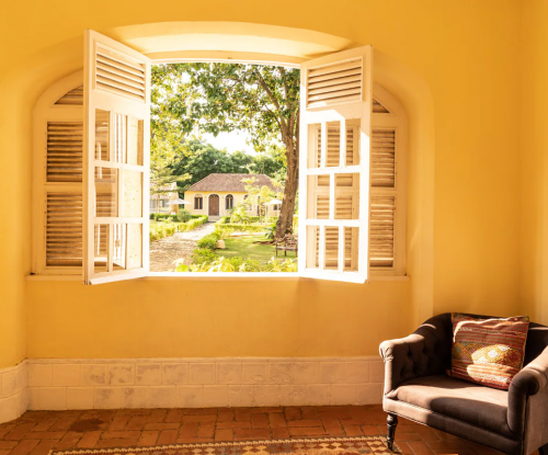 Janela iluminada no Belo Monte com vista para os jardins do pátio e edifícios amarelos, poltrona acolhedora no interior.