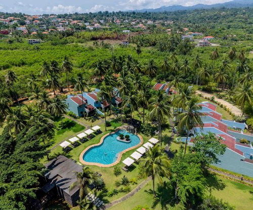 Aerial view of the Omali pool area and rooms surrounded by palm trees, with the town of São Tomé in the background