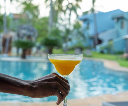 Hand holding a tropical cocktail by the pool at Omali, with palm trees and buildings in the background, São Tomé Island