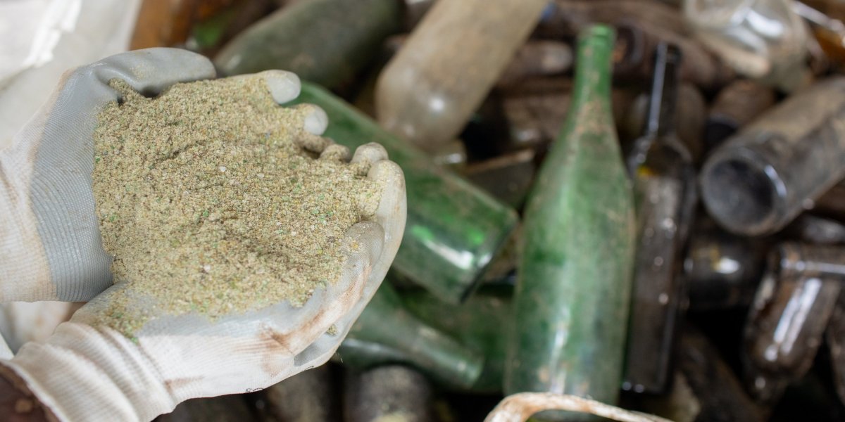 Mão de trabalhador segurando areia de vidro, com muitas garrafas de vidro vazias ao fundo.