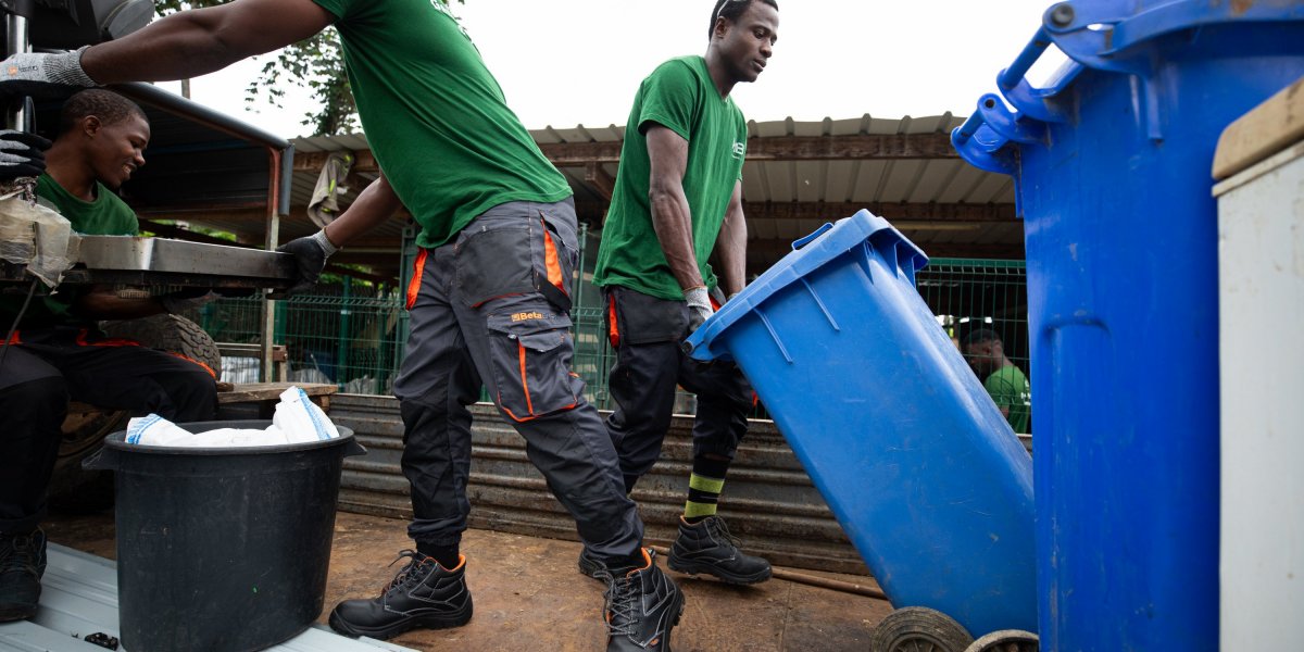 Três trabalhadores com camisetas verdes a reciclar com contentores azuis na Ilha do Príncipe.
