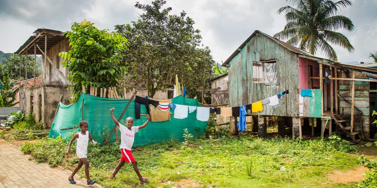 Duas crianças a rir para a câmara, com cabanas locais e roupa estendida a secar ao fundo.