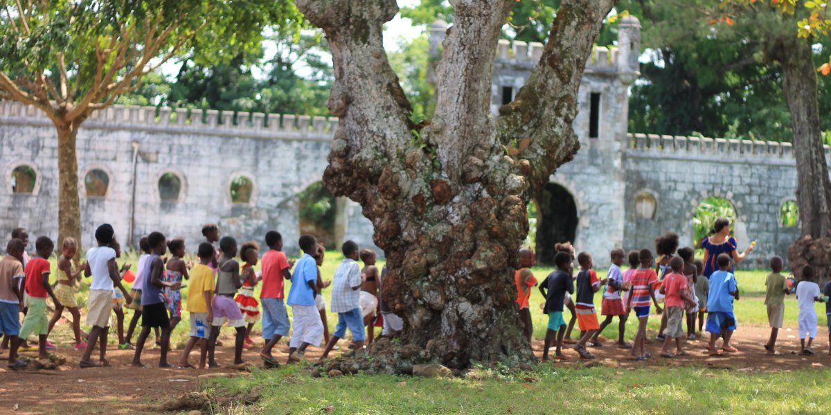 Um grupo de crianças vestidas de forma colorida atrás de uma árvore em frente à Roca Sundy, Ilha do Príncipe.