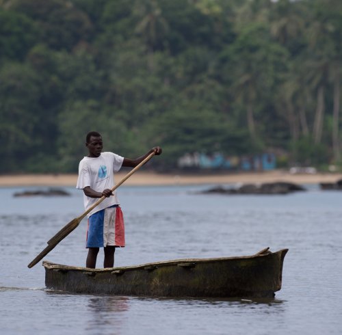 A local fisherman in colourful trousers rowing a small wooden boat, with the beach and rainforest in the background.