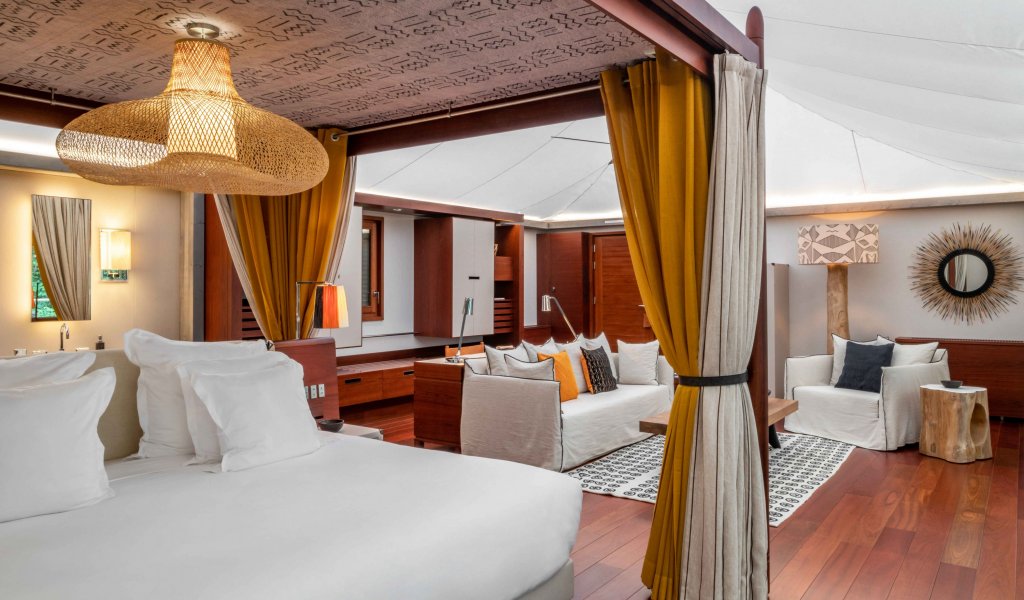 Quarto com cama de casal elegante, tapete artístico, área de estar e mesa ao fundo no Sundy Praia.