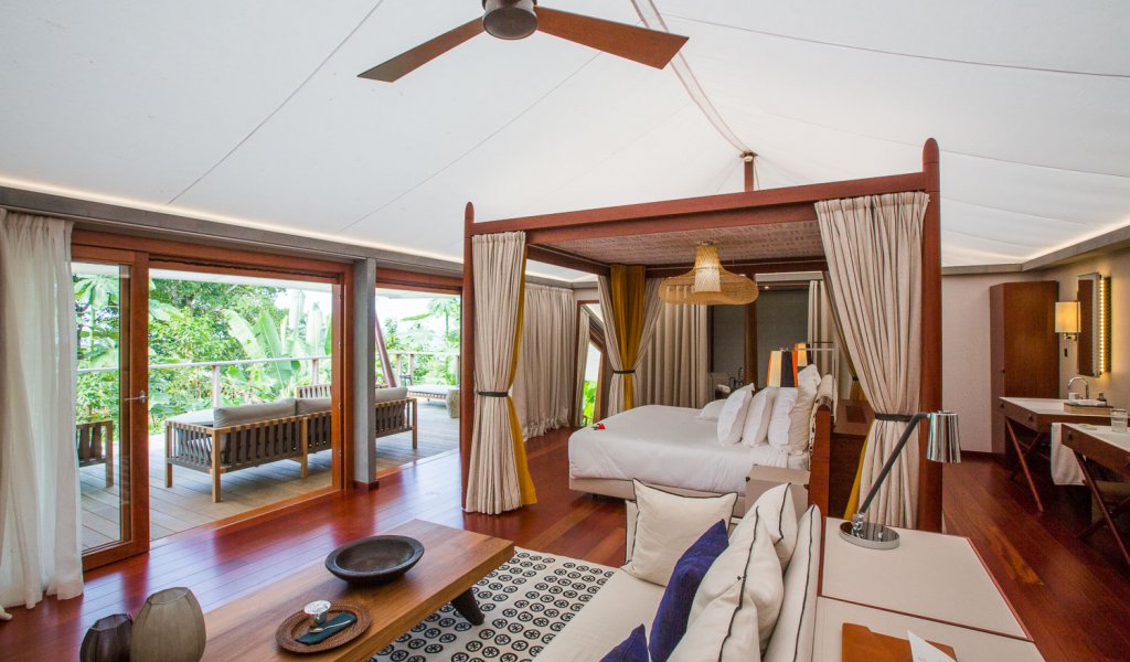 Quarto com cama de dossel elegante, tapete artístico, área de estar e mesa de madeira em primeiro plano na tenda-vila Sundy Praia.