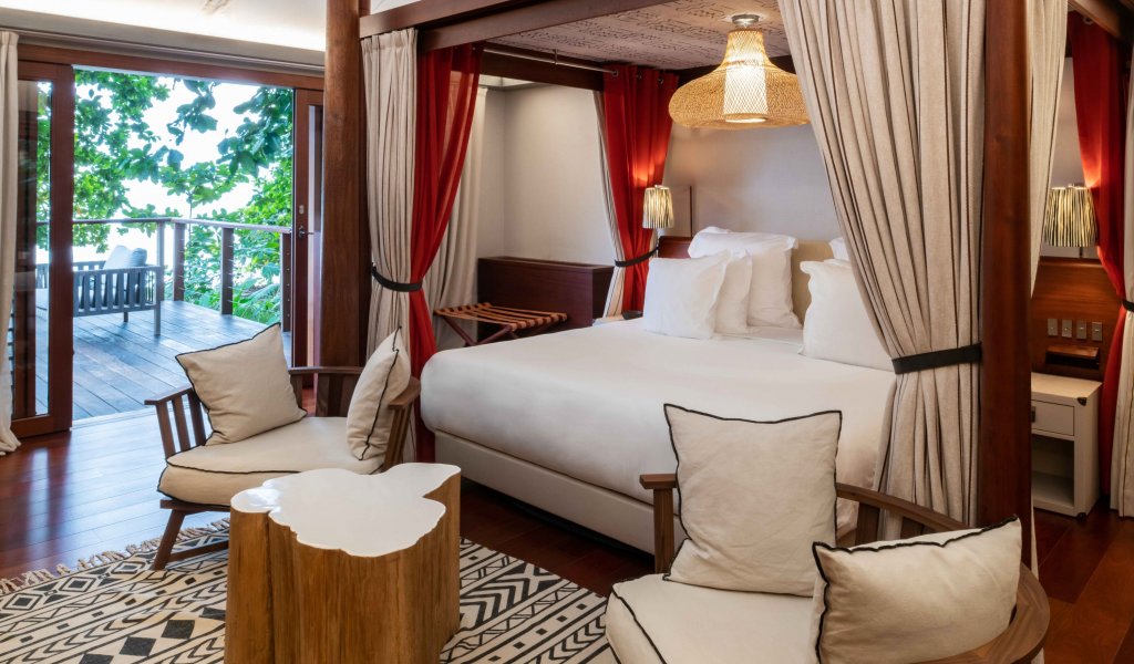 Quarto com cama de dossel elegante, tapete artístico, área de estar e mesa de madeira em primeiro plano na tenda-vila Sundy Praia.