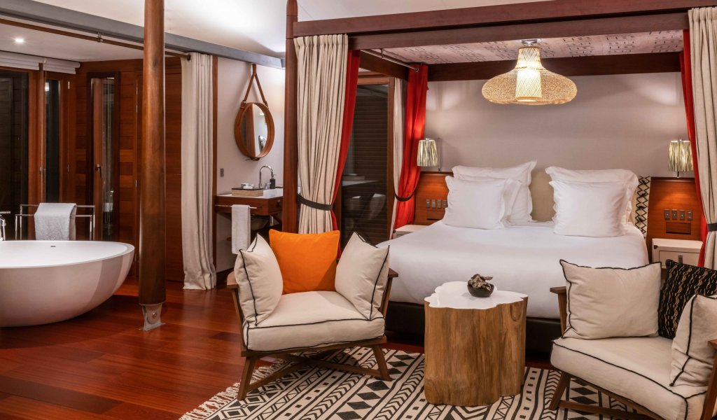Quarto com cama de dossel elegante, tapete artístico, área de estar e mesa de madeira em primeiro plano na tenda-vila Sundy Praia.